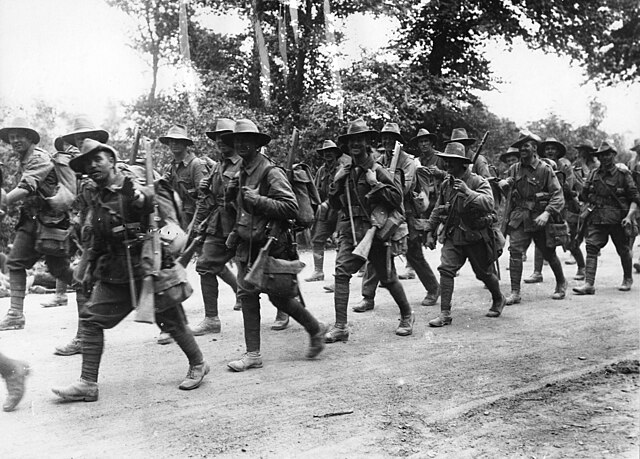 Australian WWI soldiers marching along a dirt road in France, carrying rifles and smiling, with trees in the background.