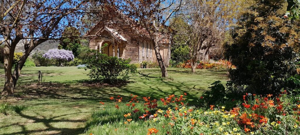 St Clements Church, Eurobin, viewed through garden trees with colourful spring flowers in the foreground and wisteria blooming in the distance.