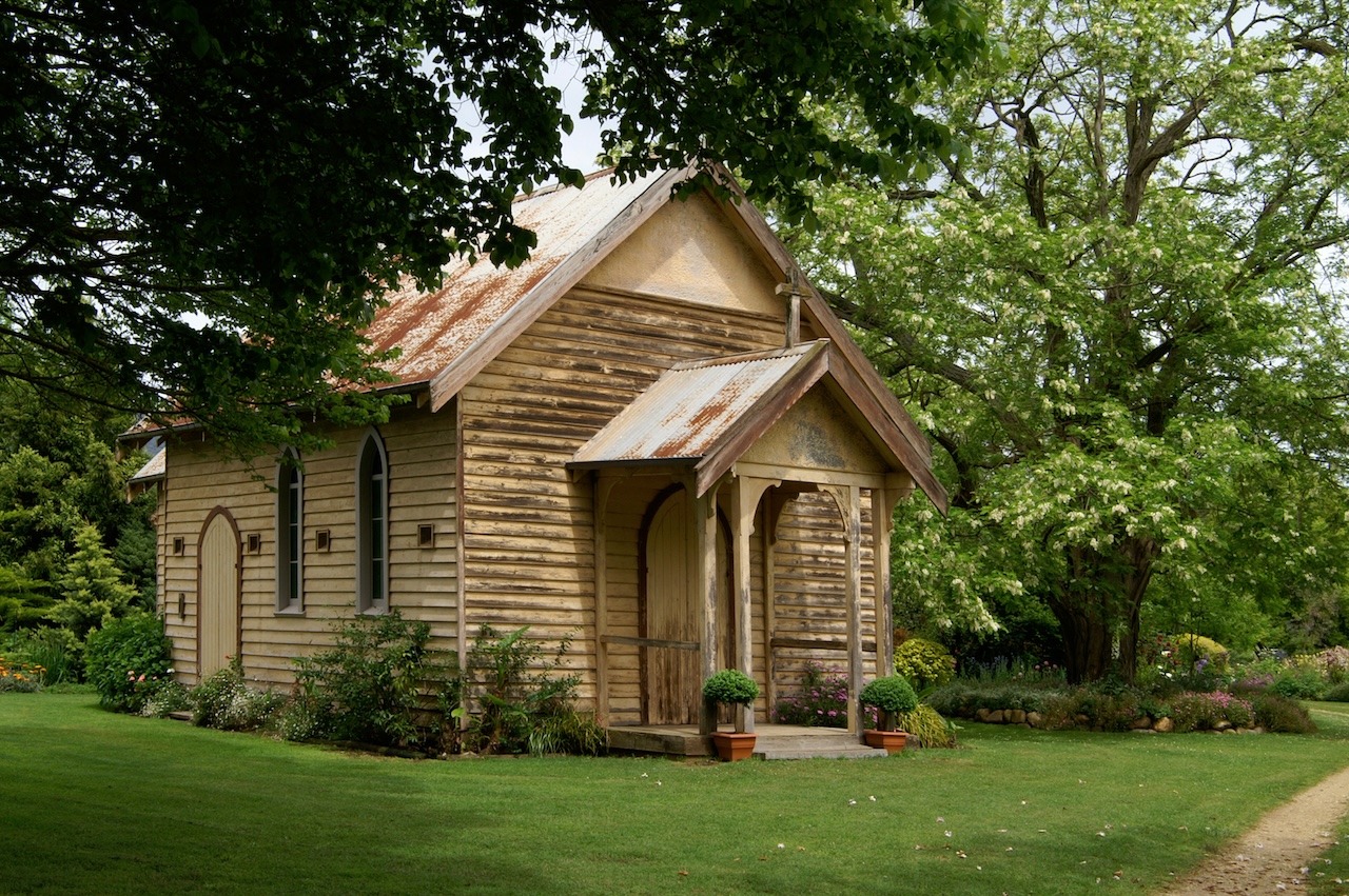 Angled view of St Clements Church, Eurobin, showing its weathered timber walls, corrugated iron roof, and arched doorway framed by lush green lawns and overhanging tree branches.