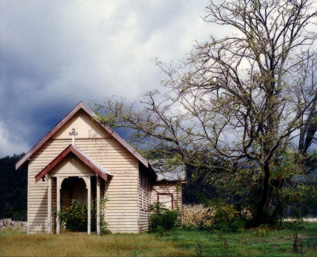 St Clements Church in Eurobin during its forgotten years - weathered timber walls, overgrown entryway, and a bare tree under a moody sky.