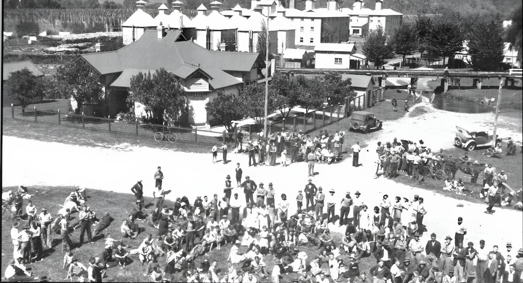 Black-and-white photograph from autumn 1929 showing a large crowd of men, women, and children gathered along a dirt roadside near Panlook’s Hop Gardens in Eurobin, Victoria. Many are seated on the grass while others stand in clusters, facing the road. In the background are buildings with steep roofs and multiple hop kilns, as well as vintage cars, bicycles, and a small bridge over a stream. The scene captures a moment of anticipation, with people waiting for seasonal work during the early years of the Great Depression.