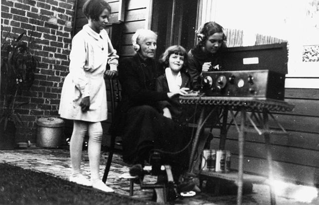 Black-and-white photograph from 1929 showing a woman seated outdoors operating a Traeger pedal wireless set, with three children gathered around watching intently. The wireless equipment is set up on a folding table beside a wooden house, with a brick wall and garden visible in the background.